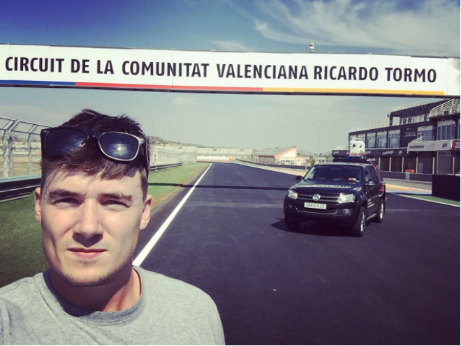 Young man on a driving race course with a black car behind him